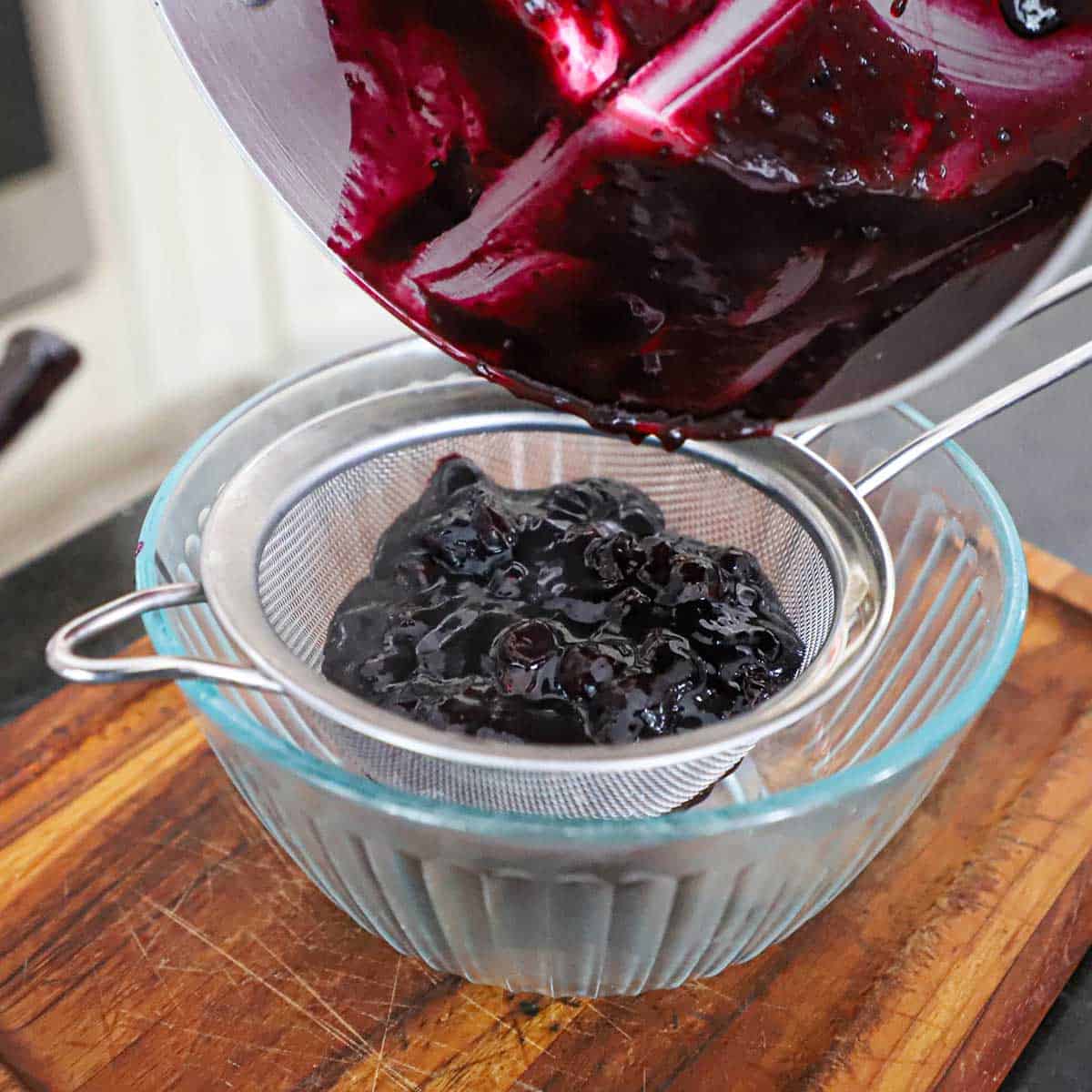 A person pouring a blueberry sauce from a sliver saucepan into a sieve that is resting over a medium-sized glass bowl on a wooden cutting board.