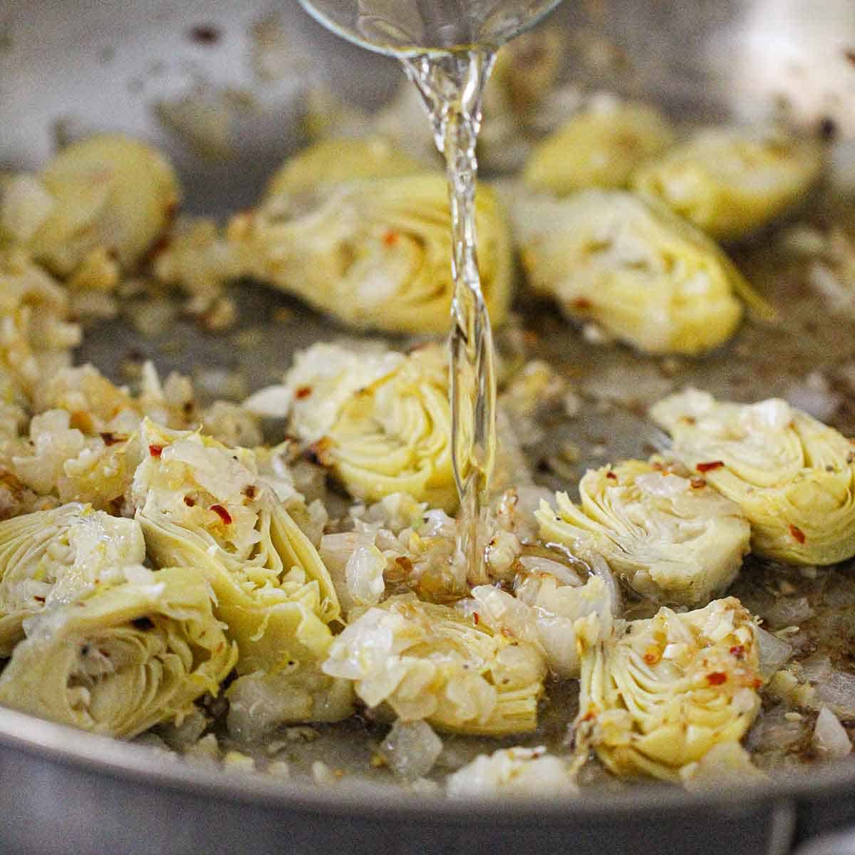 White wine being poured from a small glass into a skillet filled with simmering artichoke hearts, onion, garlic, and seasonings.