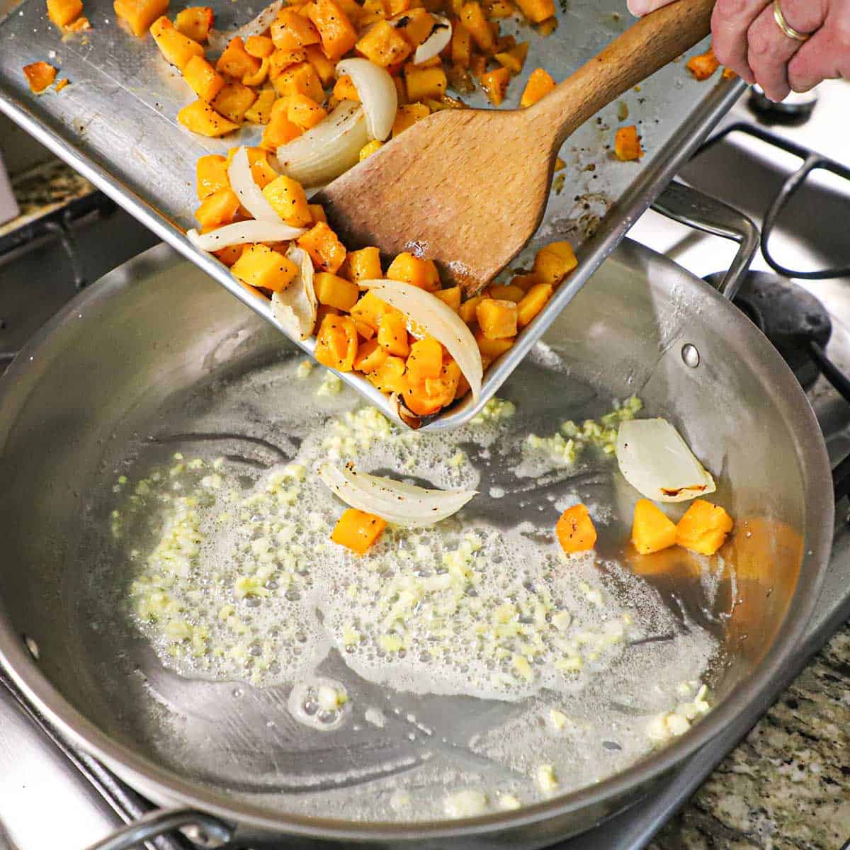 A person using a wooden spatula to transfer roasted butternut squash and onions pieces into a large skillet filled with simmering butter with minced garlic and ginger in it.