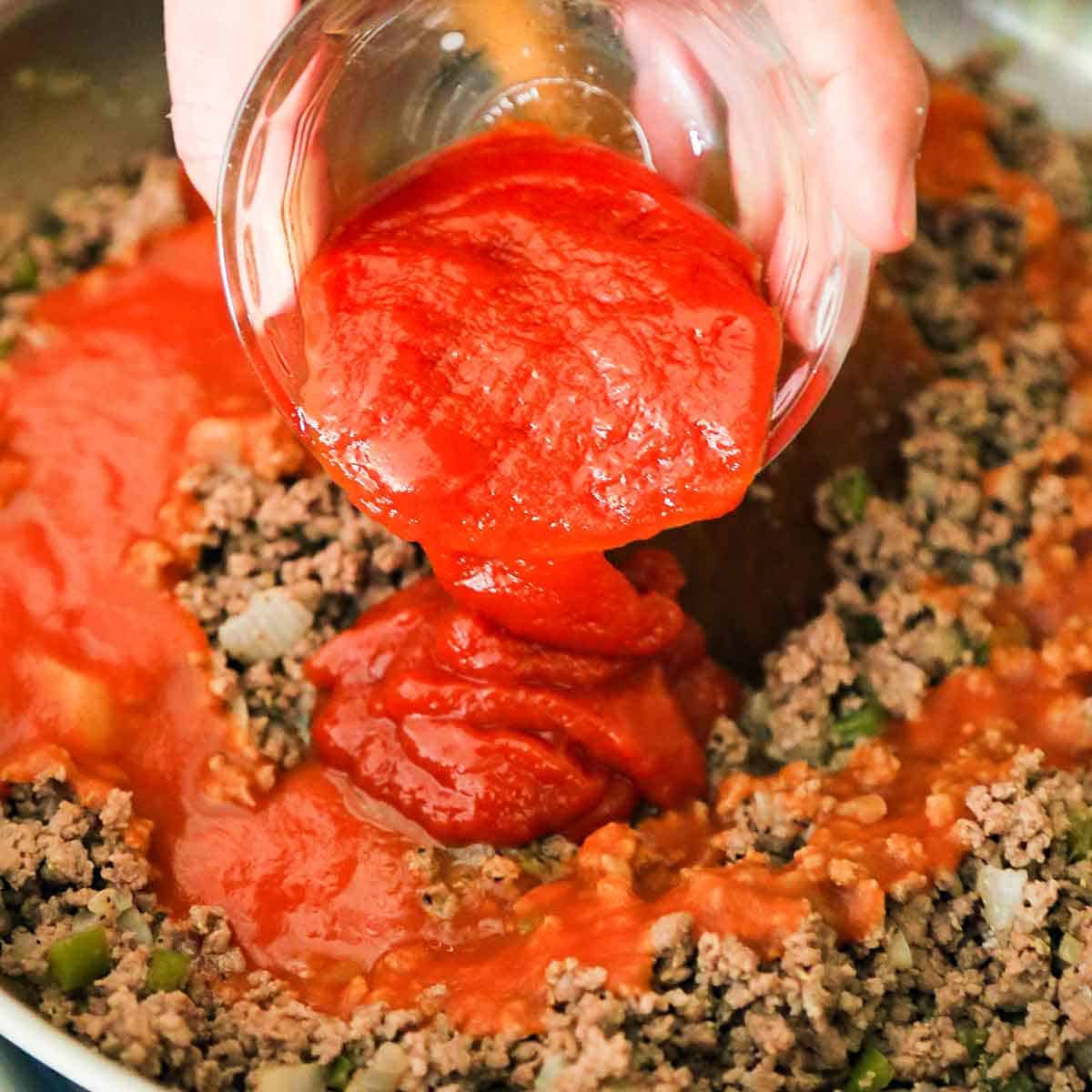 A person pouring tomato sauce from a small glass bowl into a skillet filled with cooked ground beef, chili sauce, and sautéed vegetables.