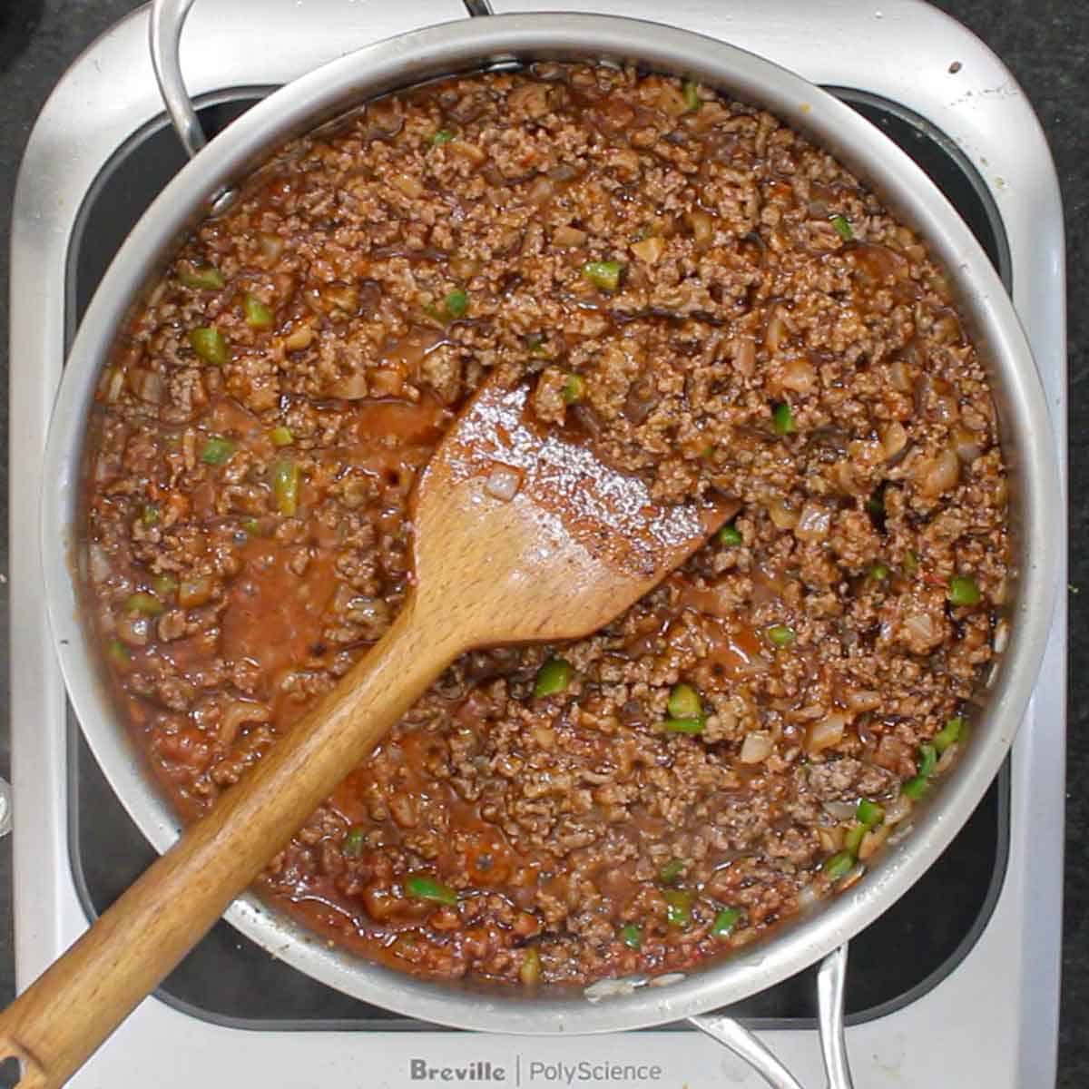 A person using a large wooden spatula to stir a sloppy Joe meat mixture in a large silver skillet.