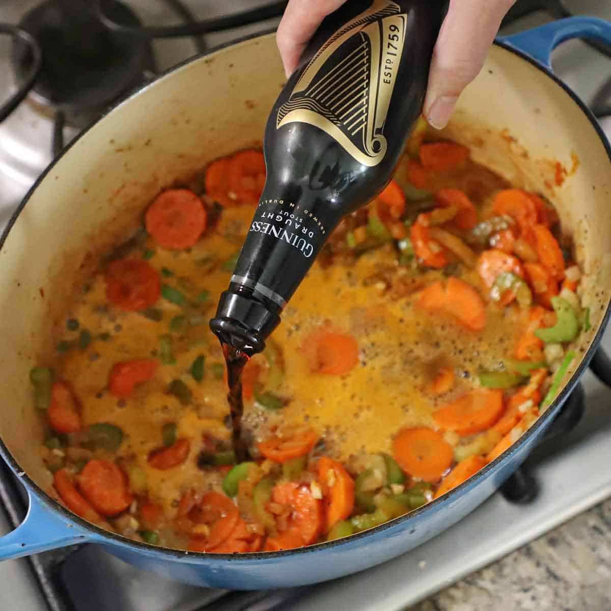 A person pouring stout beer from a Guinness bottle into a large Dutch oven that is filled with simmering carrots, onions, and celery.