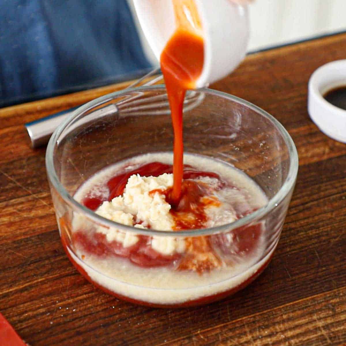 A person pouring Tabasco hot sauce from a small white bowl into a larger glass bowl filled with the unmixed ingredients for homemade cocktail sauce.