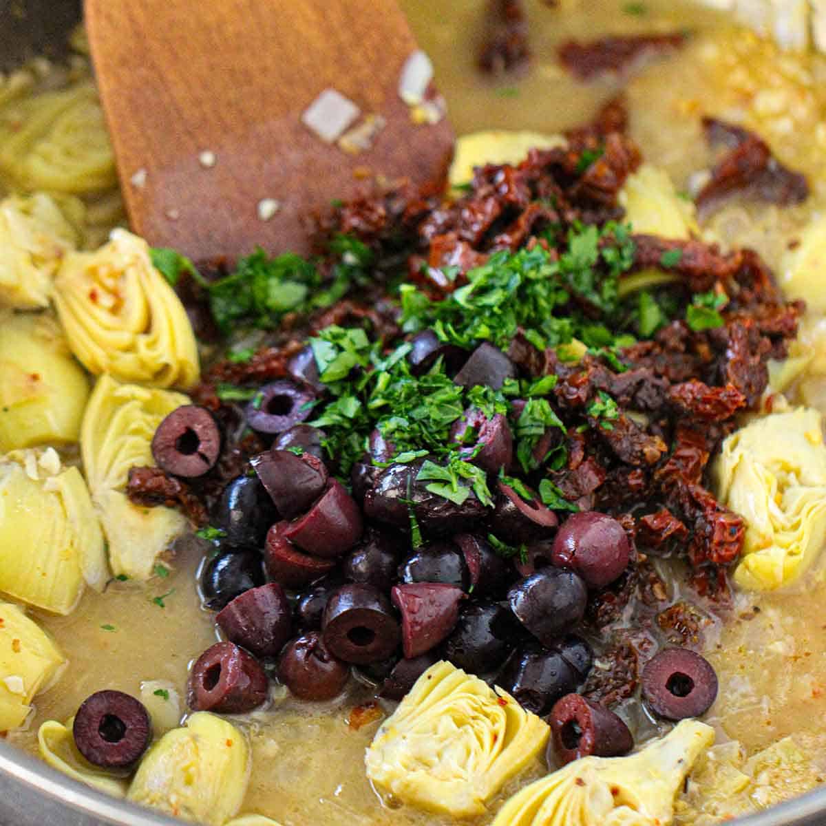 A wooden spatula being used to stir chopped kalamata olives, sun-dried tomatoes, and fresh herbs into sautéed artichoke hearts that are simmering in a white wine and chicken stock sauce.