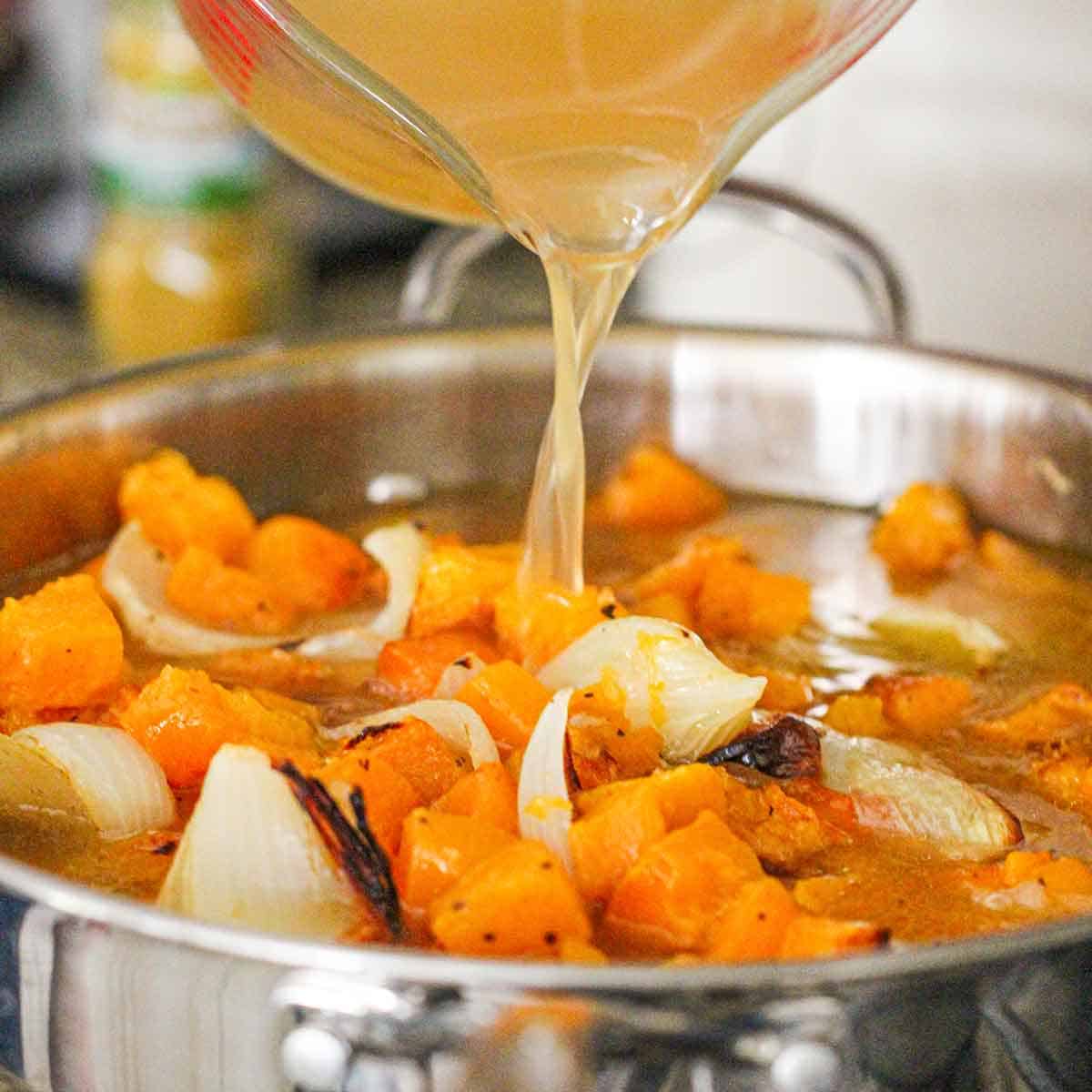 A person pouring chicken stock from a large glass measuring cup into a large skillet filled with roasted butternut squash and onions.