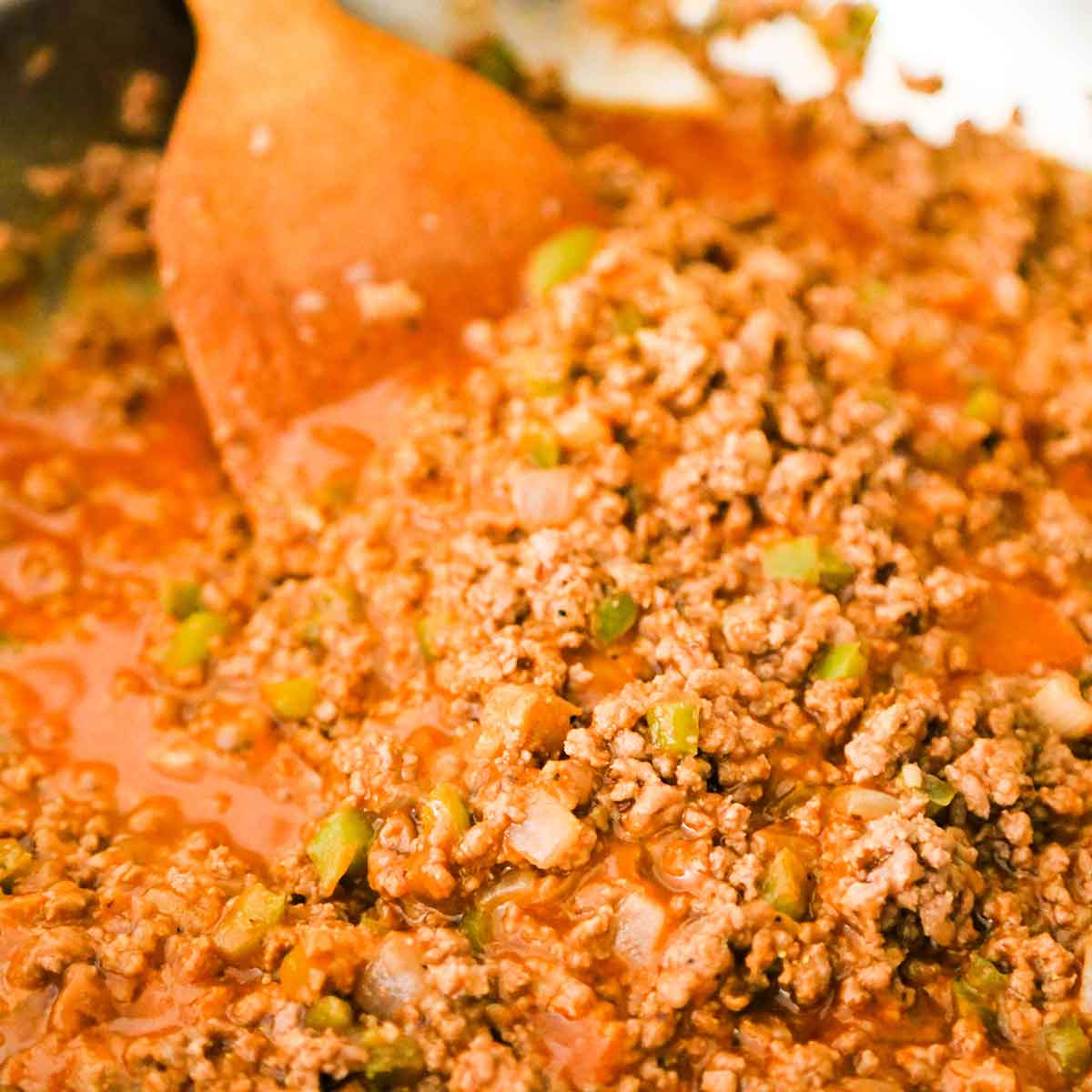 A person using a wooden spatula to stir a saucy mixture of homemade Sloppy Joes filling in a large skillet.