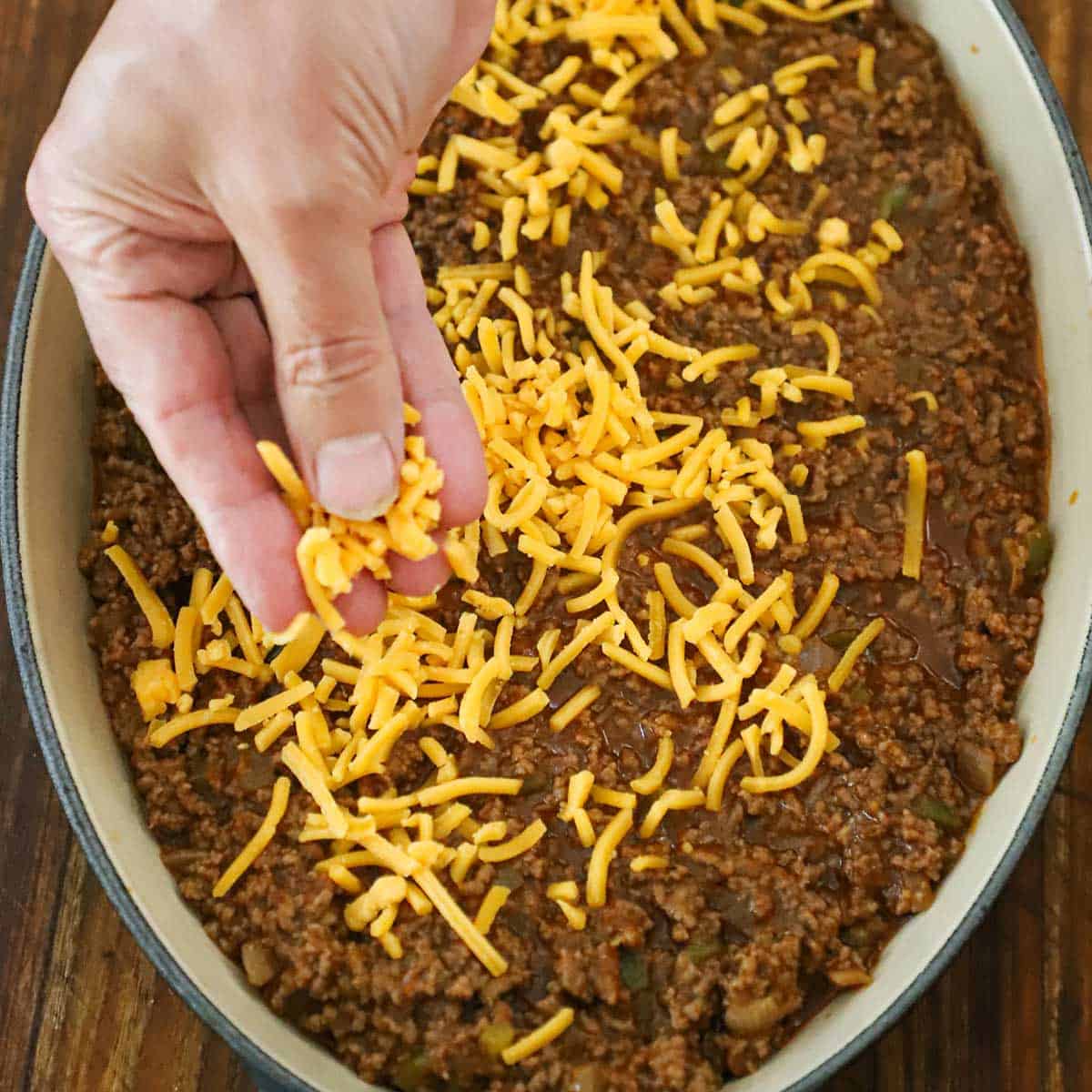 A person sprinkling shredded cheddar cheese over a sloppy Joe meat mixture that is in an oval ceramic baking dish.
