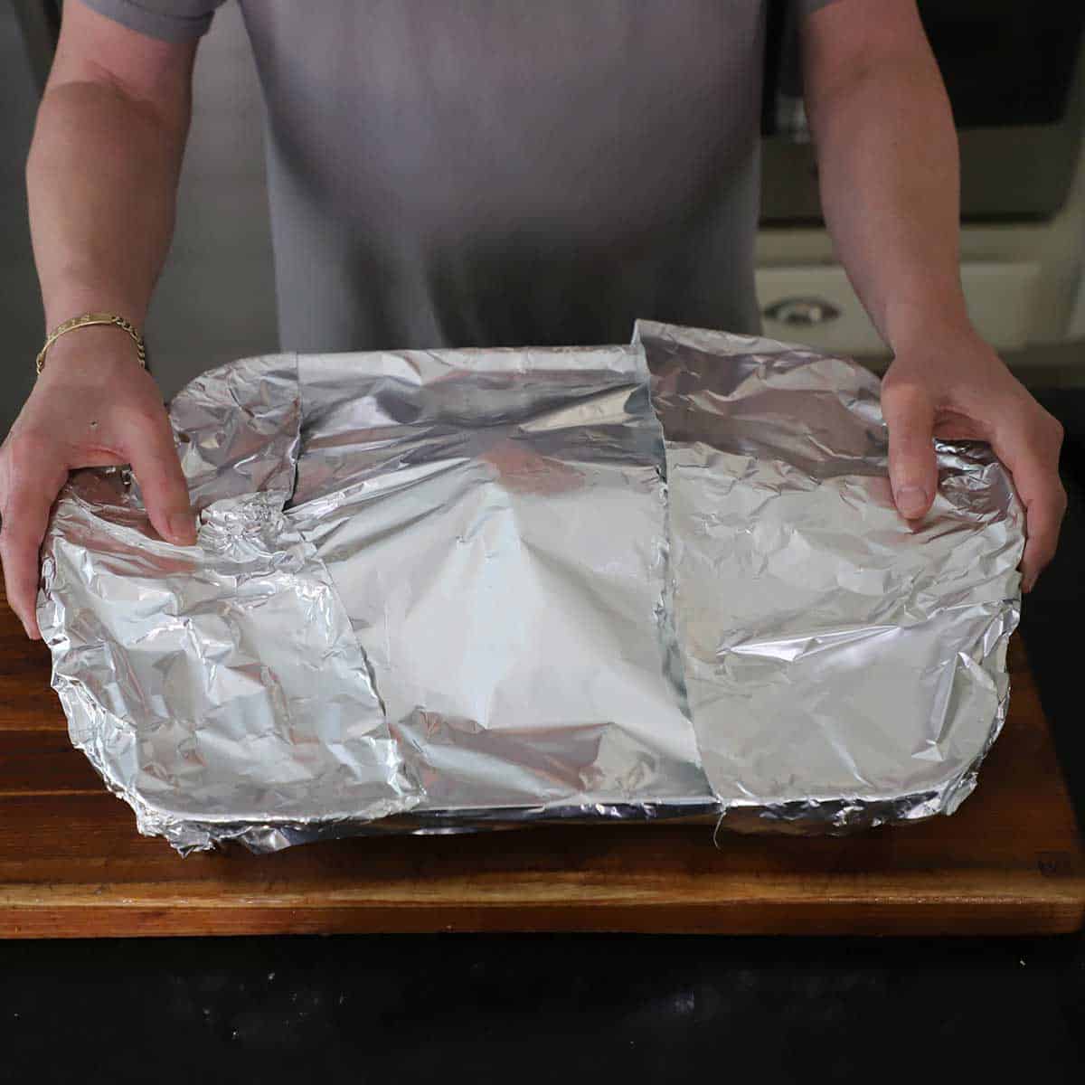 A person pressing down heavy duty foil over a large roasting pan on a wooden cutting board.