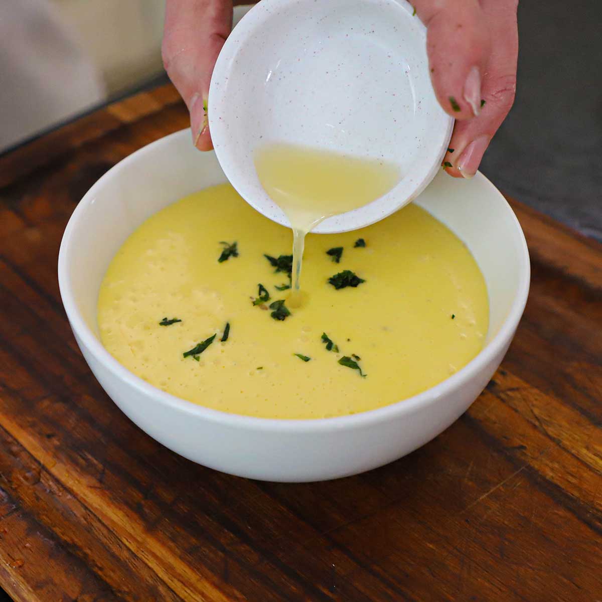 A person pouring fresh lemon juice from a small bowl into a larger white bowl holding Blender Béarnaise Sauce garnished with chopped tarragon.
