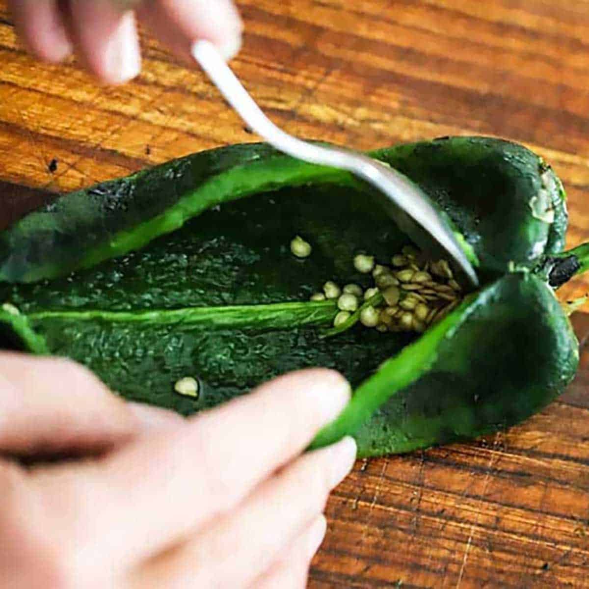 A person using a spoon to scrape seed out of the inside of an opened roasted poblano chile.