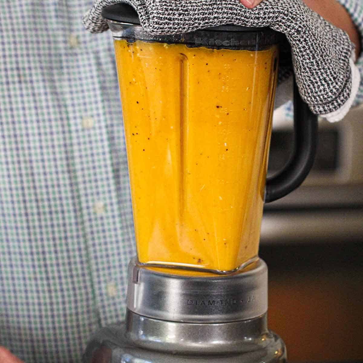 A person holding a kitchen towel over the top of a blender that is filled to the top with puréed roasted butternut squash soup.
