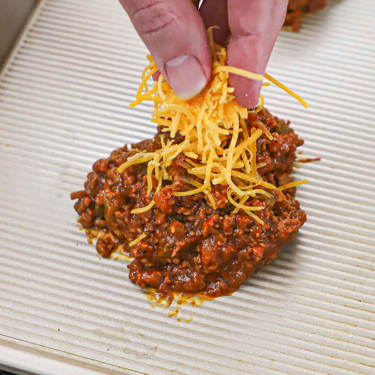 A person placing a mound of shredded cheddar cheese on a pile of a homemade sloppy Joes mixture on a silver baking pan.