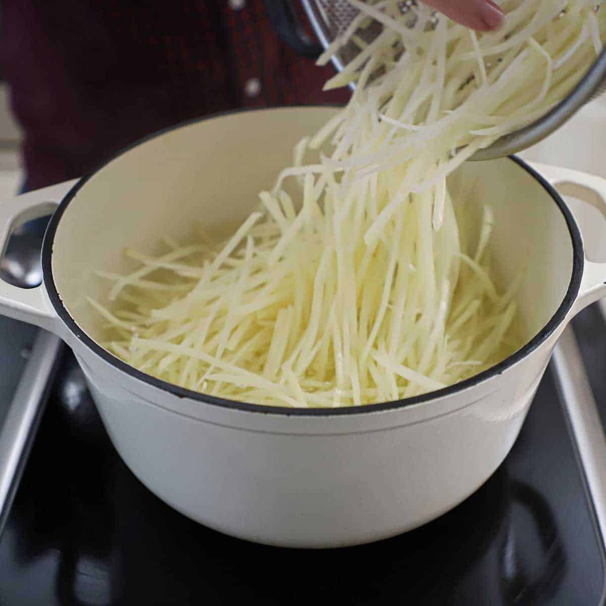A person transferring uncooked shoestring potatoes into a bowl of boiling water with vinegar and salt.