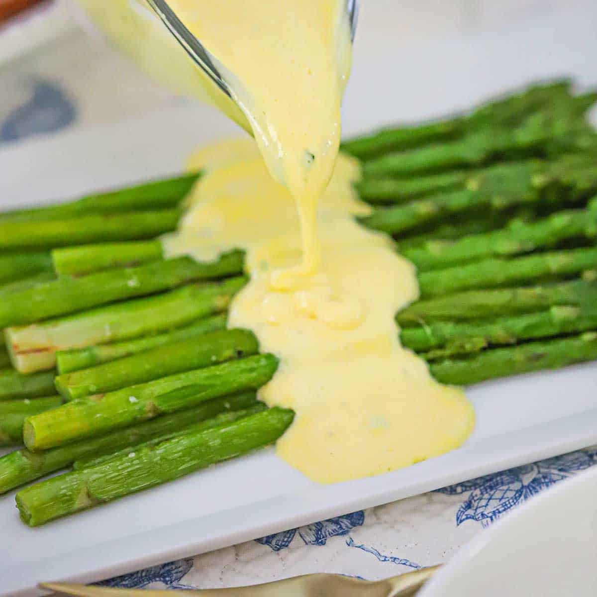 A person pouring blender Béarnaise sauce from a glass gravy boat onto a pile of pan-seared asparagus on a white rectangular platter.