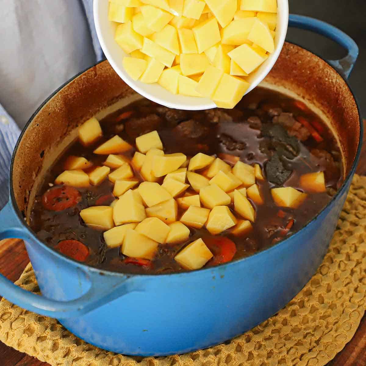 A person transferring cubed gold potatoes from a white bowl into a large pot of simmering classic Guinness beef stew.