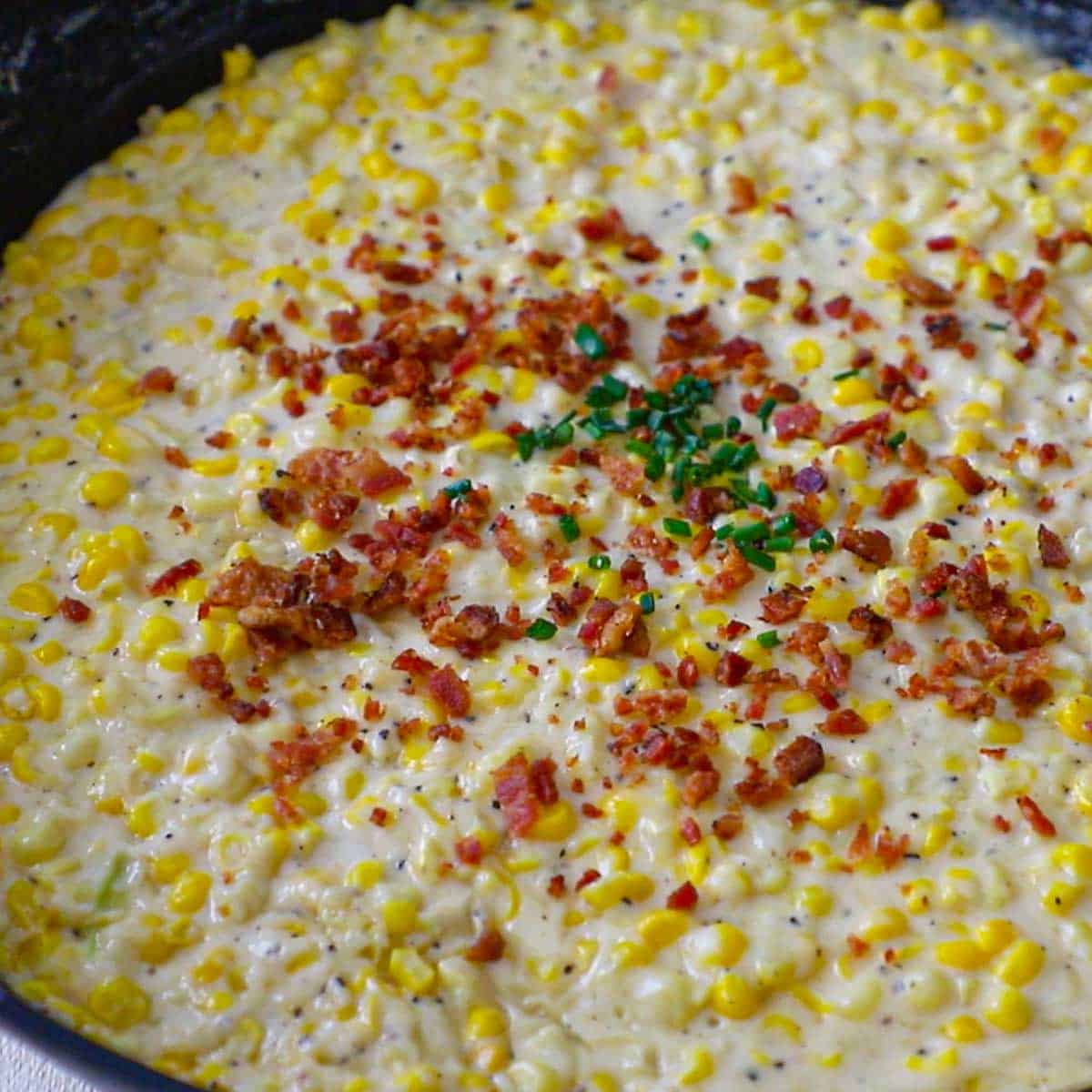 A close-up view of a cast-iron skillet filled with freshly prepared homemade creamed corn topped with crispy bacon bits and snipped chives.