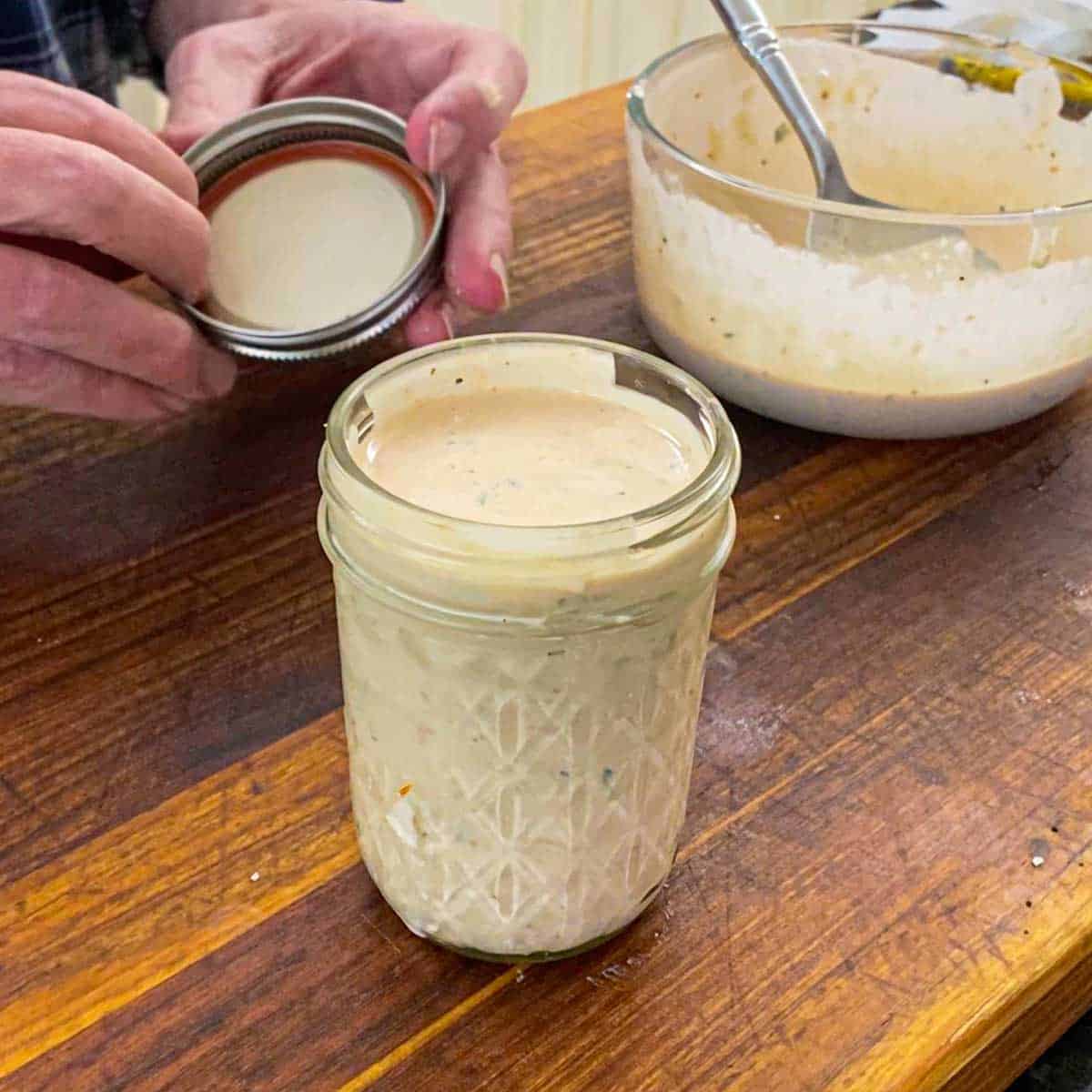 A person holding a jar lid near the jar that is filled to the top with homemade Russian dressing on a wooden cutting board.