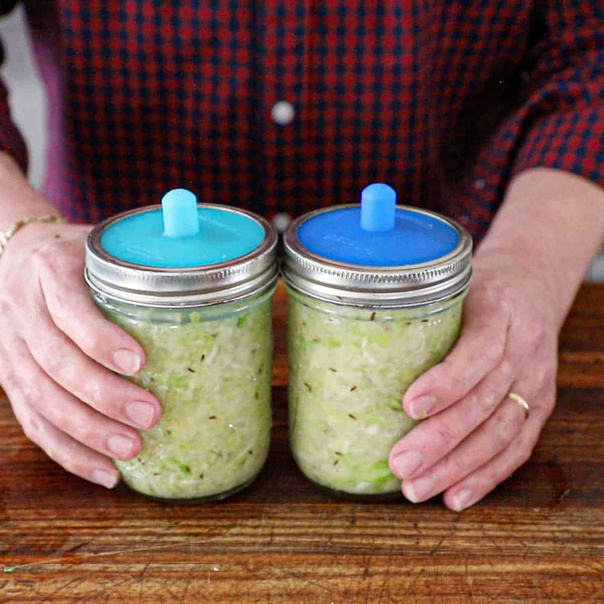 A person gripping two glass jars filled with green cabbage and topped with a "pickle pipe" on a wooden cutting board.