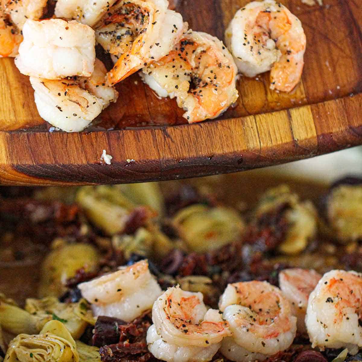 Cooked pink shrimp being transferred from a small wooden cutting board into a large skillet filled with a simmering Mediterranean pasta sauce.