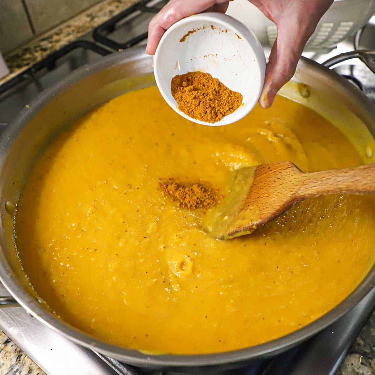 A person adding curry powder from a small white bowl into a large skillet that is filled with simmering roasted butternut squash soup.