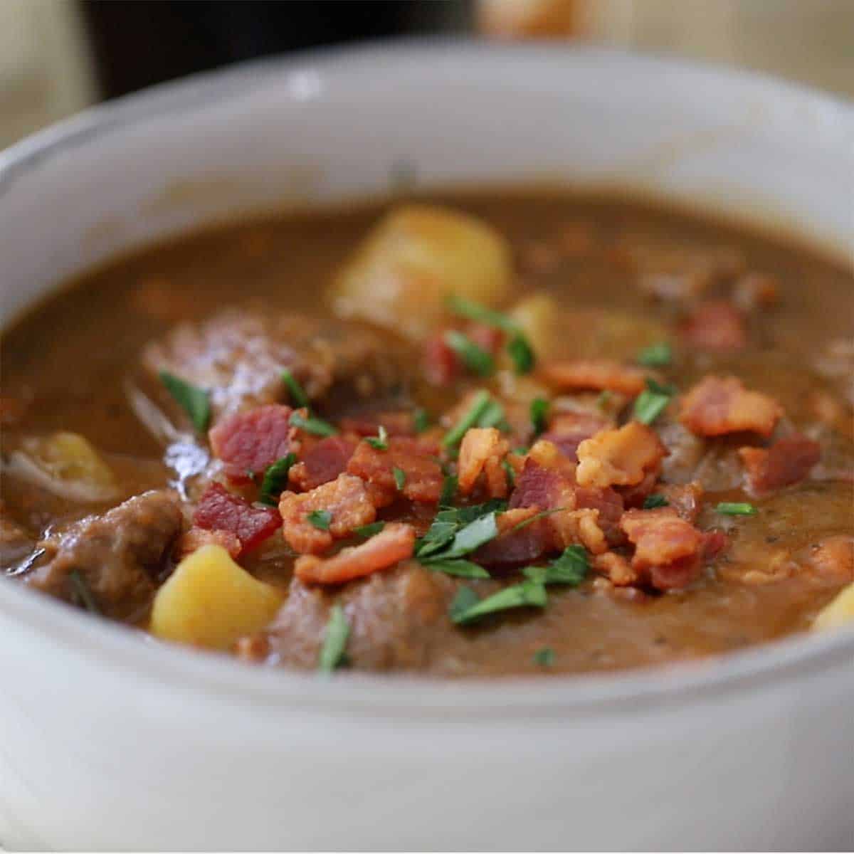 A close up view of a bowl filled with classic Guinness beef stew and topped with crispy pieces of bacon and chopped Italian parsley.