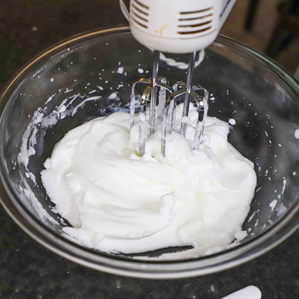 A person using a hand-held electric mixer to beat egg whites into soft peaks in a glass bowl.