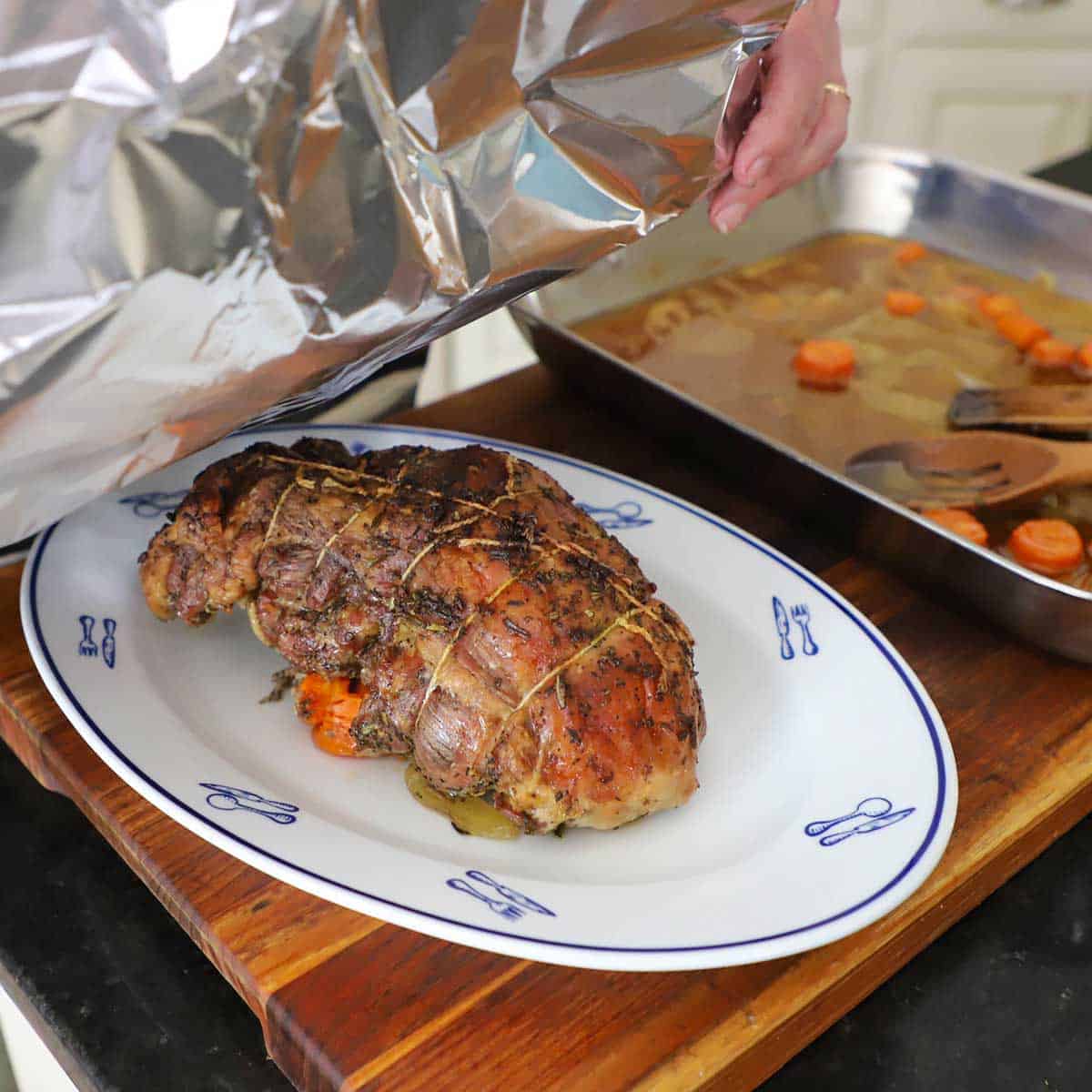 A person tenting a slow-roasted lamb shoulder on a platter with a large piece of heavy duty foil.