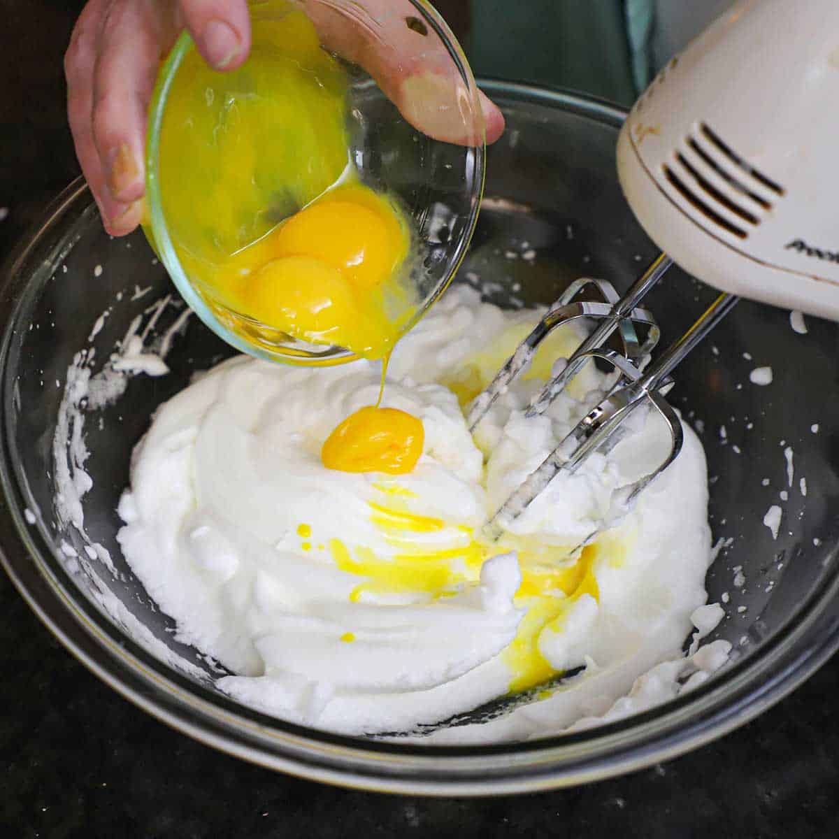 A person transferring three egg yolks from a small glass bowl into a bowl that is filled with egg whites that have been whipped into soft peaks.