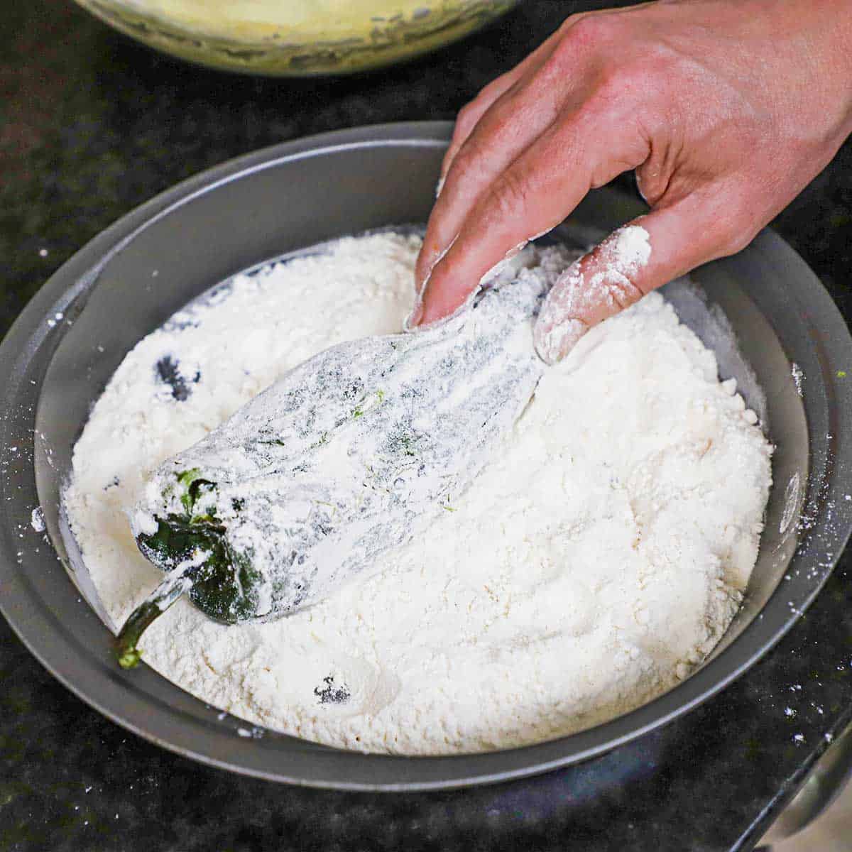 A person dredging a stuffed roasted chile in flour that is in a circular cake pan.