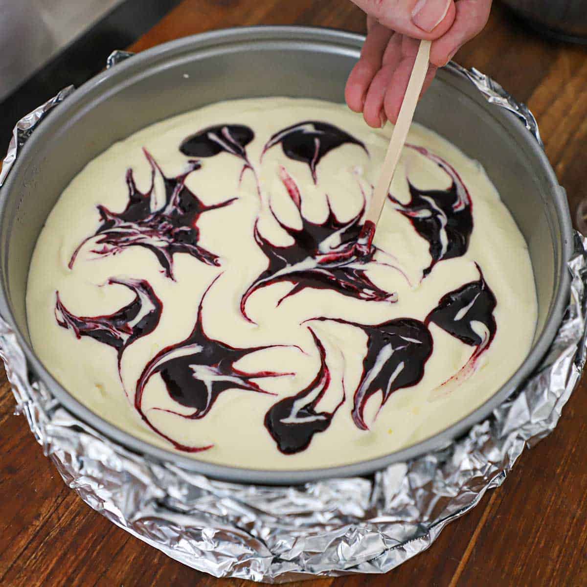 A person using a wooden coffee stirrer to make swooping designs from dollops of a blueberry sauce on top of an uncooked lemon blueberry cheesecake in a springform pan.