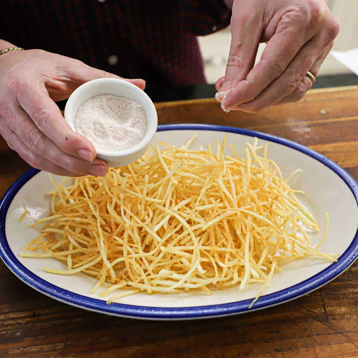 A person sprinkling fine sea salt over a pile of shoestring French fries on a large platter sitting on a wooden cutting board.