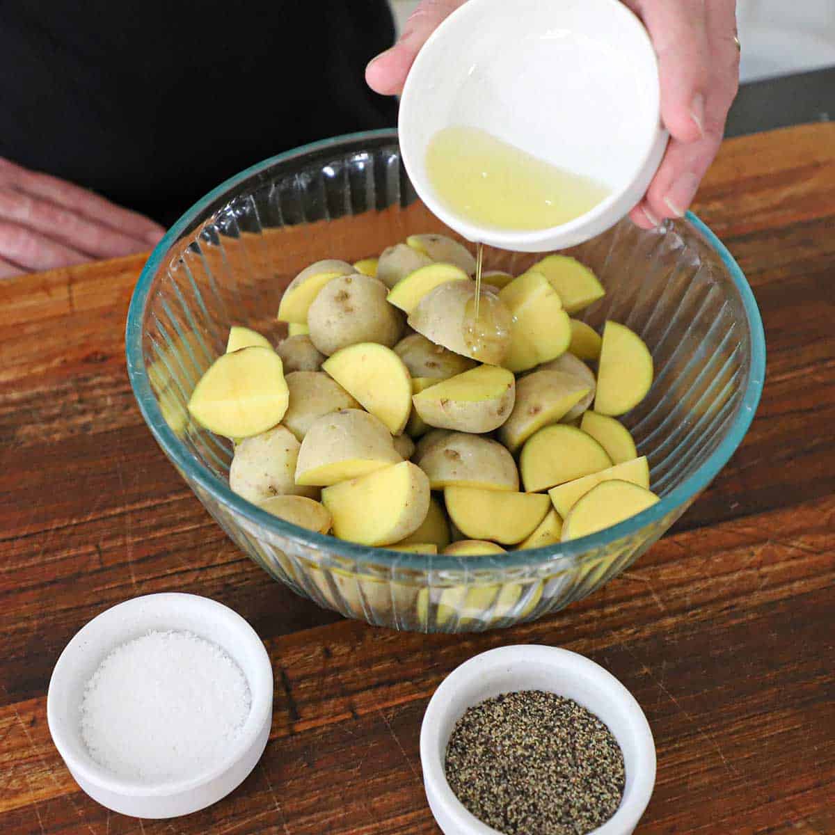 A person pouring olive oil from a small white bowl into a glass bowl filled with baby gold potatoes that have been halved.