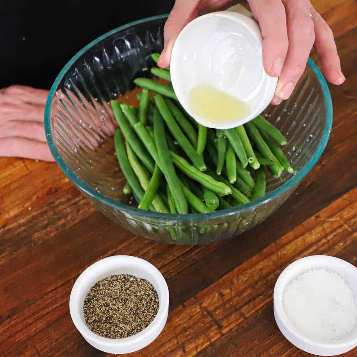 A person pouring olive oil over fresh green beans that are in a medium glass bowl on a wooden cutting board.
