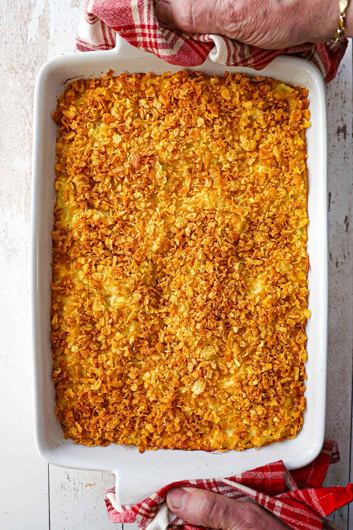 An overhead view of a person using two red-checkered linens to hold the handles of a white baking dish containing an untouched hashbrown casserole from scratch.