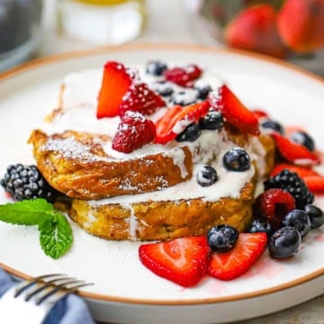 A large white dinner plate filled with two caramelized brioche French toast slices that are topped with a mascarpone cream sauce and macerated berries.