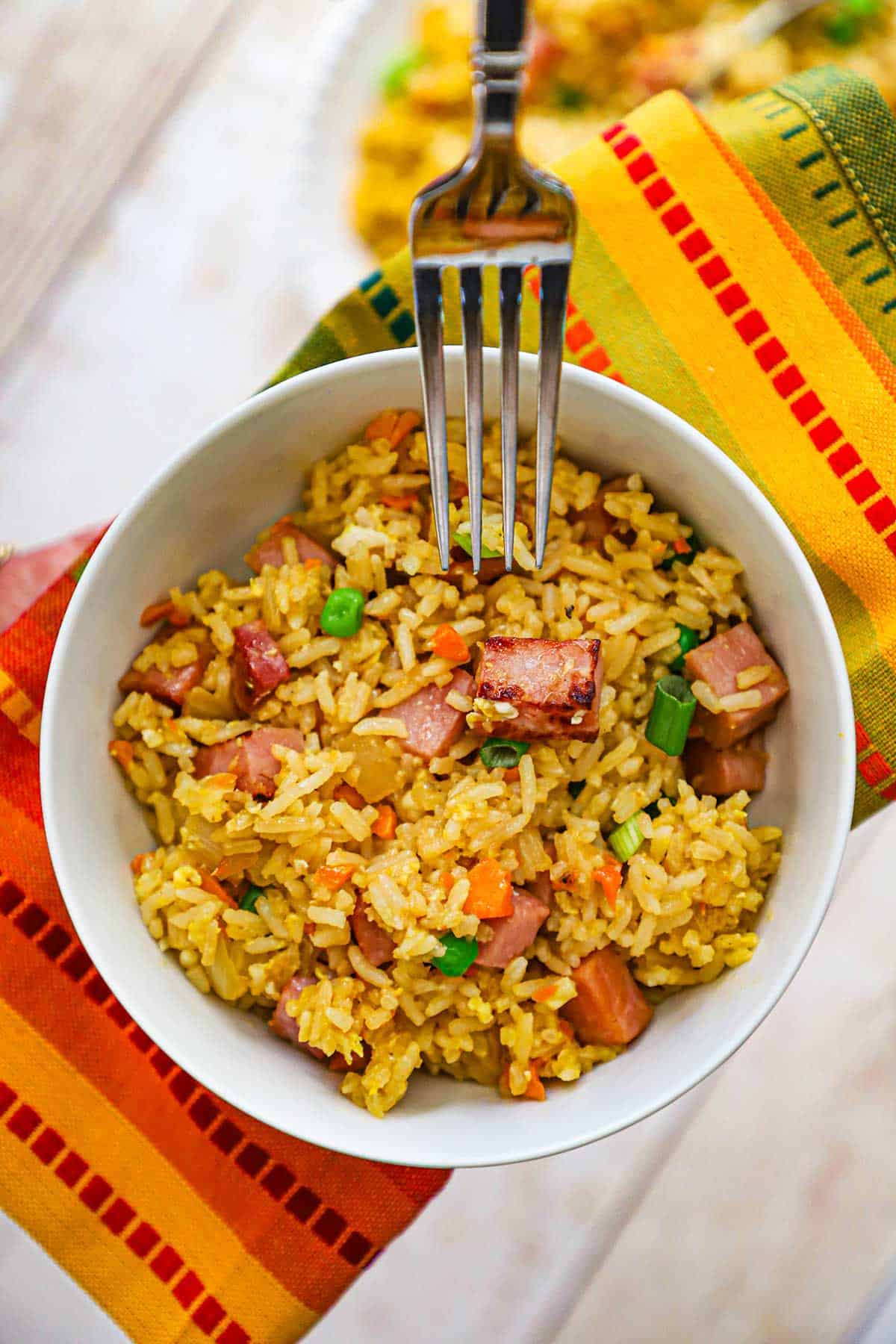 A person holding a fork over a white bowl filled with a serving of leftover ham fried rice with a colorful napkin underneath the bowl.