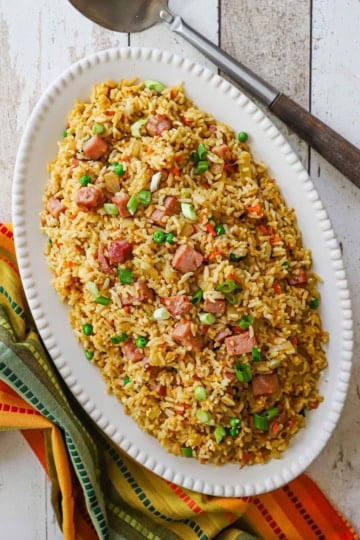 An overhead view of an oval platter containing a large serving of leftover ham fried rice with a colorful napkin and a large serving spoon nearby.