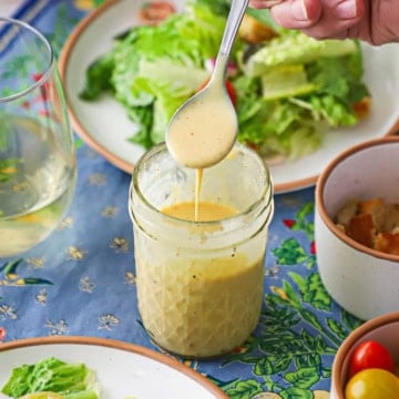 A person holding a spoonful of Restaurant-Style Honey Mustard Dressing over a glass jar holding the same in front of a side salad on a salad plate.