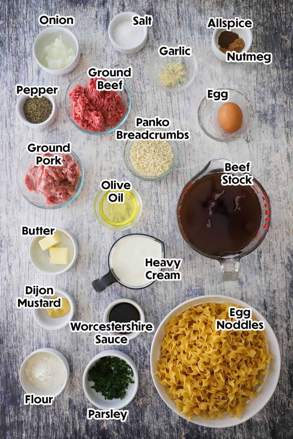 An arrangement of ingredients for Swedish meatball pasta on a grey wooden background including ground beef, ground pork, breadcrumbs, an egg, grated onion, butter, beef stock, cream, egg noodles, and seasonings.