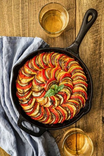 An overhead view of a cast-iron skillet ratatouille with two glasses of white wine sitting nearby as well as a light blue napkin.