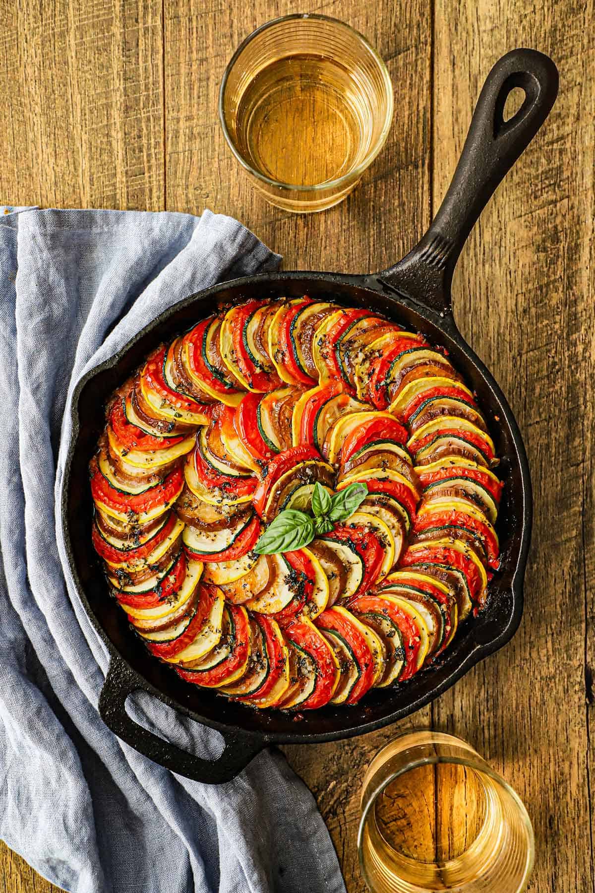 An overhead view of a cast-iron skillet ratatouille with two glasses of white wine sitting nearby as well as a light blue napkin.