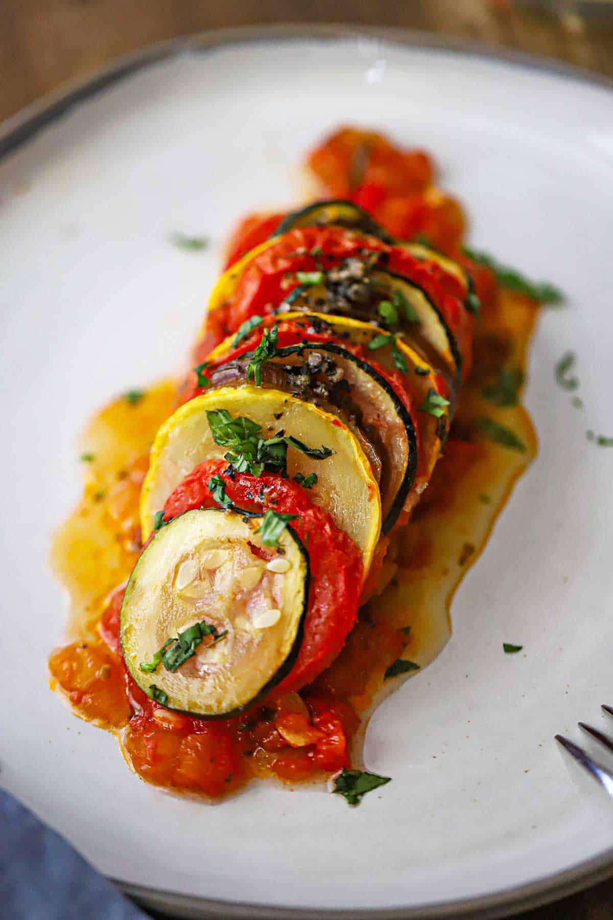 A slanted row of thinly sliced vegetables from a cast-iron skillet ratatouille resting on a thin bed of sautéed tomatoes and red bell pepper all on a white dinner plate.