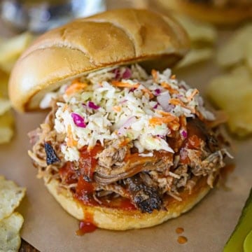 A close-up view of a smoked pulled pork sandwich resting on brown sandwich paper with shredded pork, BBQ sauce, and coleslaw all nestled between two toasted hamburger buns.