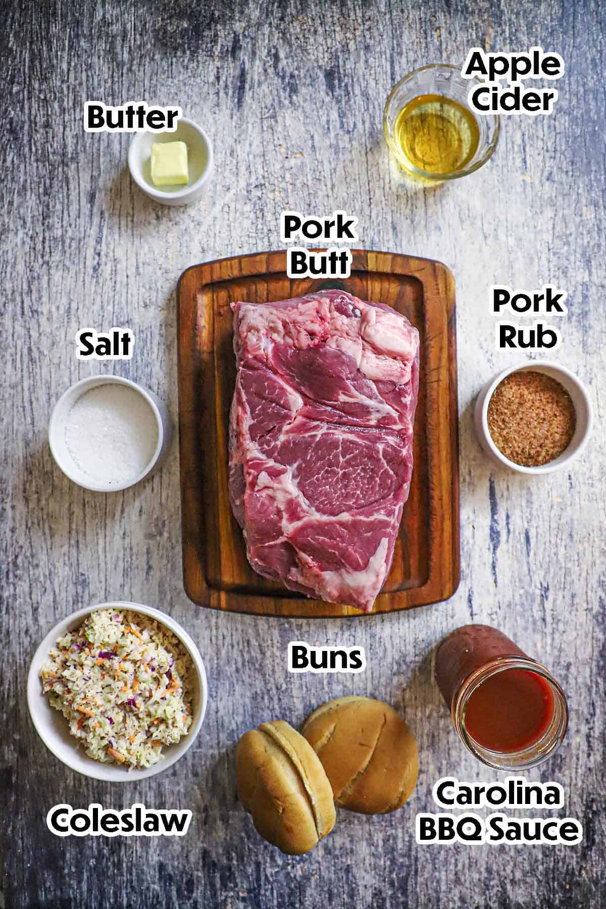 An arrangement of ingredients for smoked pulled pork including a pork butt, pork rub, salt, apple cider, butter, coleslaw, hamburger buns, and bbq sauce.