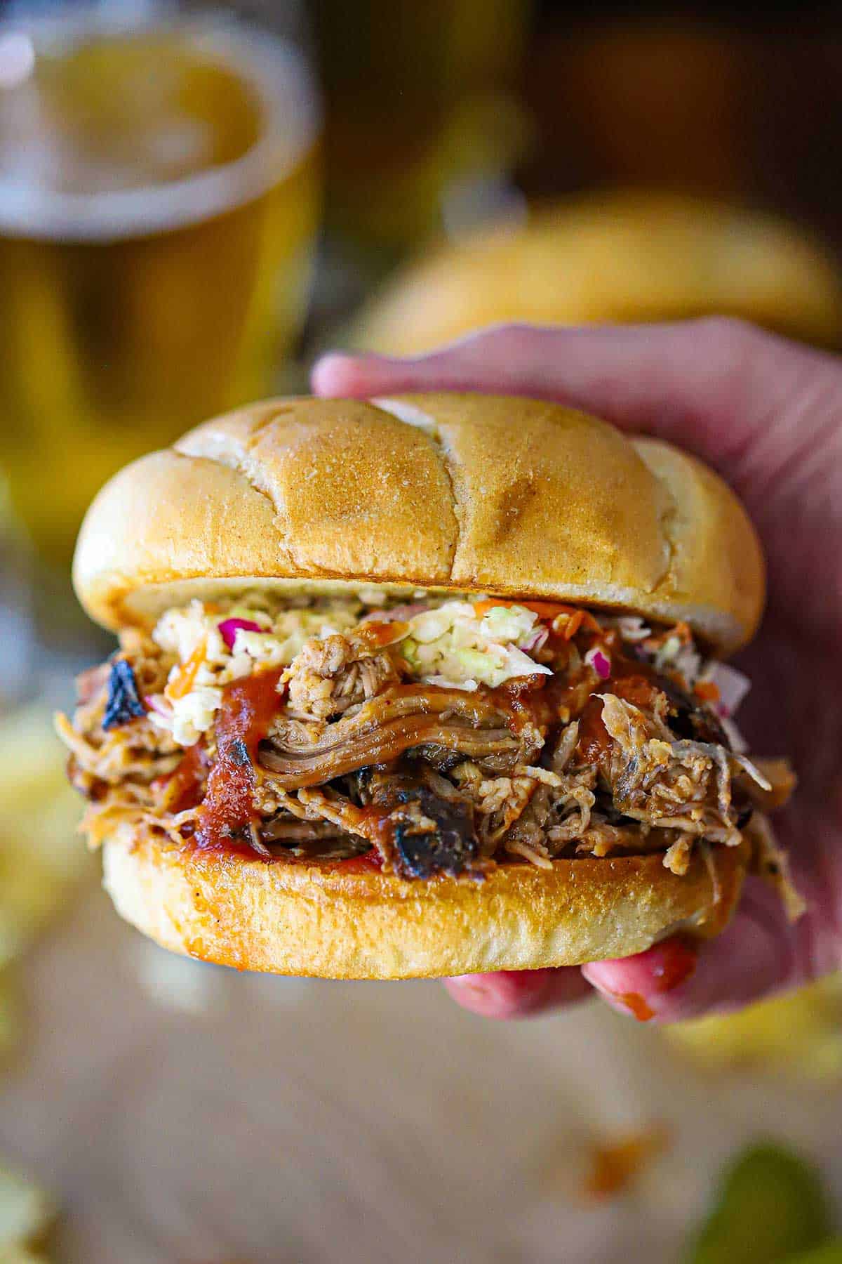 A person holding a smoked pulled pork sandwich with one hand revealing the shredded pork, BBQ sauce, and coleslaw on toasted buns.