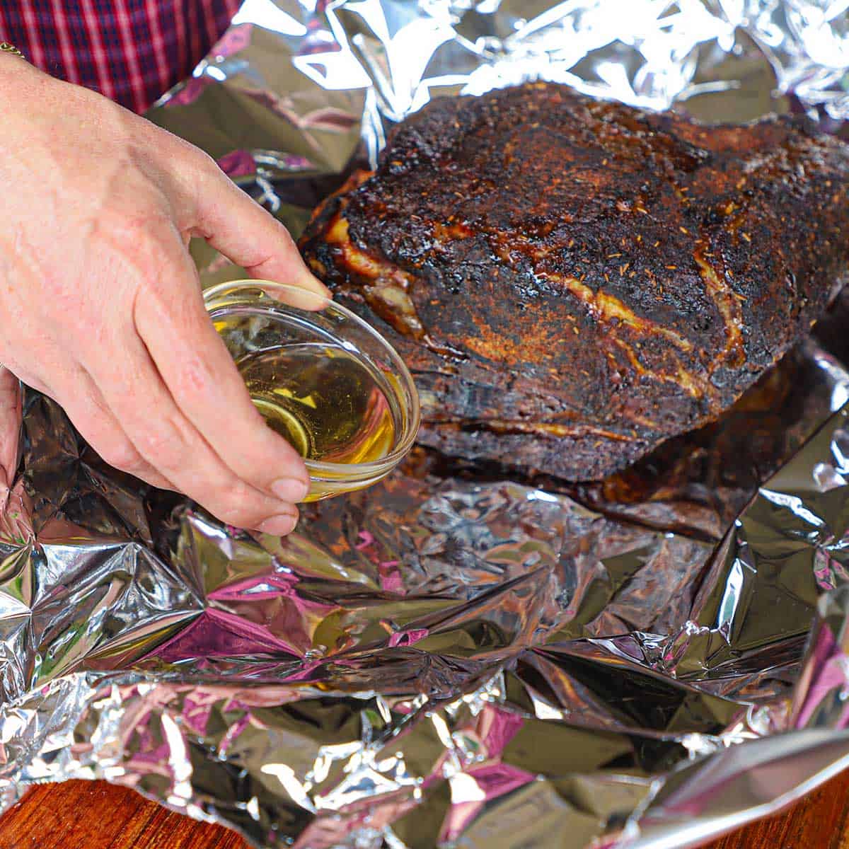 A person transferring apple cider from a small glass bowl around the base of a smoked pork butt that is sitting on two large pieces of heavy-duty foil on a wooden cutting board.