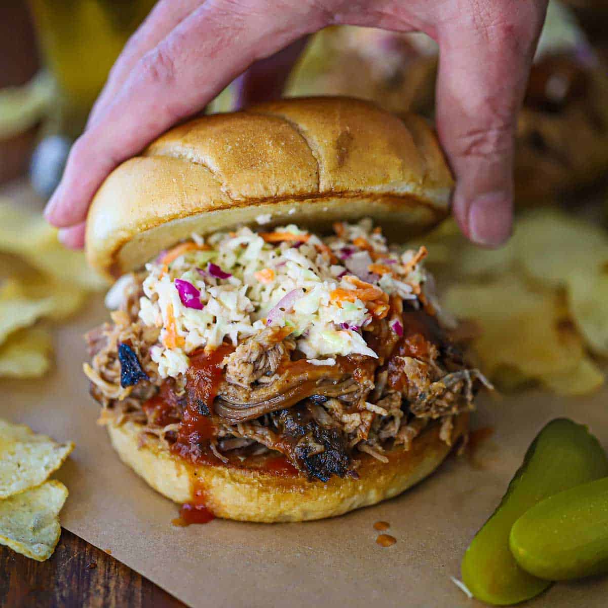 A person placing a top toasted hamburger bun onto a smoked pulled pork sandwich topped with BBQ sauce and homemade coleslaw.