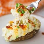 A person using a fork to lift the filling out of a twice baked potato that is resting on a white salad plate.
