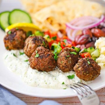 A close-up view of five Greek Meatballs resting on a bed of homemade tzatziki sauce surrounded by a cucumber salad, crumbled feta cheese, olives, red onion, and pita triangles.
