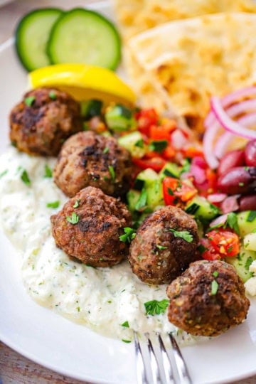 Five Greek Meatballs resting on a bed of homemade tzatziki sauce surrounded by a cucumber salad, crumbled feta cheese, olives, red onion, and pita triangles all on a white dinner plate.